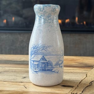 May include: A ceramic milk bottle vase with a speckled white finish and a blue rim. The front features a detailed blue illustration of a water mill, a house, and bare trees. The design has a smooth, rounded neck and a wide base for stability.