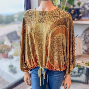May include: A long-sleeved, patterned top in shades of brown, orange, and yellow, with a drawstring waist. The top features an intricate, repeating design and is paired with blue jeans. The sleeves are gathered at the wrists.