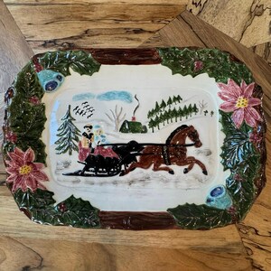 May include: A rectangular ceramic serving platter featuring a raised relief winter scene of a brown horse pulling a black sleigh with two figures. The border is decorated with green holly leaves, red berries, and pink poinsettia flowers.