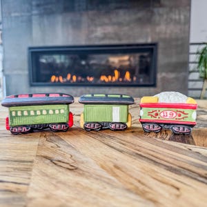 May include: Three decorative ceramic train cars in a row. The train cars are painted in shades of green, red, and yellow with black accents. The train cars are sitting on a wooden surface.