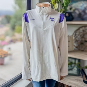 May include: White long-sleeve athletic jacket with a high collar and a partial zipper. Features purple shoulder accents, a small Nike logo, and a purple "T" logo. The jacket is made of a lightweight material.