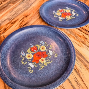 May include: Two blue ceramic plates with a floral design. The plates have a white background with red, yellow, and green flowers. The flowers are painted in a folk art style.