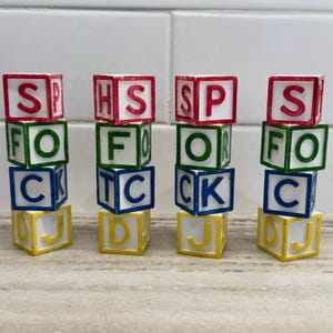 May include: Four vertical stacks of four square alphabet blocks each. The blocks feature red, green, blue, and yellow borders with corresponding colored letters. The letters visible on the faces include S, P, H, F, O, T, C, K, D, and J.
