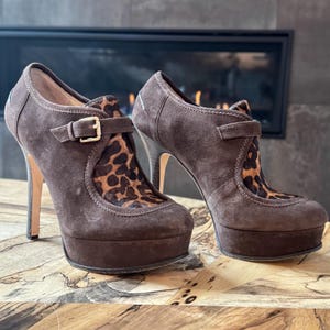 May include: A pair of brown suede platform heels with a leopard print accent on the vamp. The shoes feature a buckle closure and a high stiletto heel. The platform measures approximately 1 inch.