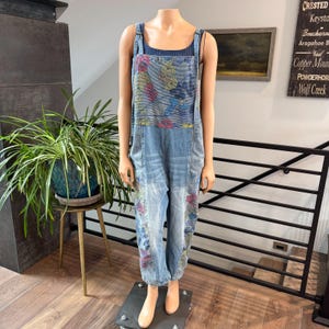 May include: Light blue denim jumpsuit with floral embroidery. The jumpsuit features adjustable straps, a bib front, and a relaxed fit. Floral patterns in pink, yellow, and blue adorn the bib and legs. The model is standing on a gray platform.