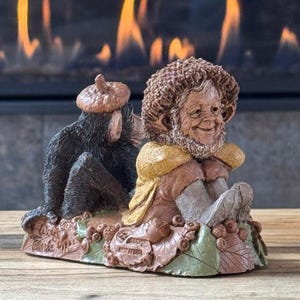 May include: A brown and tan figurine featuring a small creature with a textured cap and yellow shawl sitting among sculpted leaves and acorns, accompanied by a dark brown monkey-like figure wearing an acorn cap, with the text Wolfe Clarke 1994.