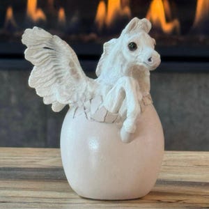May include: A white figurine of a winged horse emerging from a cracked egg. The creature has detailed feathers on its wings, a textured mane, and dark eyes. The egg is smooth and rounded, with visible cracks around the top where the horse appears.