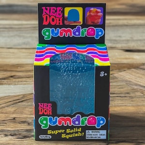 Pode incluir: Um brinquedo Nee Doh Gumdrop numa caixa. A caixa é preta com detalhes coloridos e as palavras "Nee Doh" e "gumdrop" em uma fonte divertida. A caixa também diz "Super Solid Squish!" e tem um aviso para peças pequenas.