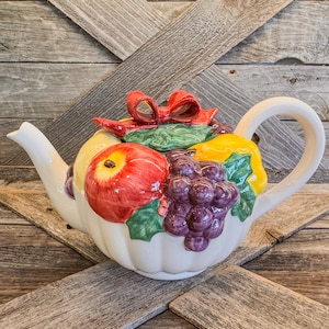 May include: A white ceramic teapot decorated with a fruit motif. The teapot features a red apple, purple grapes, a yellow pear, and green leaves. A red bow sits on top of the teapot.