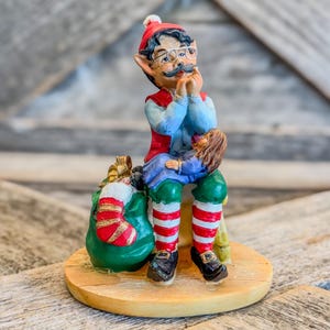 May include: A whimsical Christmas figurine of an elf wearing a red hat, vest, and striped socks, seated with a child on his lap. The elf has a mustache and glasses. A green sack with a red and gold stocking is beside him. The figurine is on a wooden base.