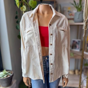 May include: Cream-colored corduroy shirt jacket with a button-down front, two chest pockets, and long sleeves. The jacket is displayed on a mannequin, revealing a red top underneath. The mannequin is wearing blue jeans.