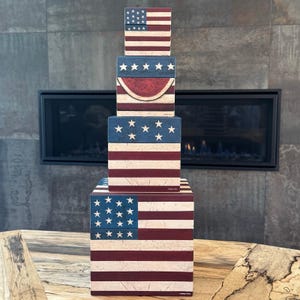 May include: A stack of four square wooden boxes featuring a distressed red, white, and blue patriotic theme. The boxes display American flag patterns, white stars on blue fields, and a central red watermelon slice graphic on the second box from top.