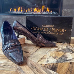 May include: A pair of brown snakeskin-patterned pumps with a pointed toe and small heel. The shoes are next to a black box with gold text that reads "COUTURE DONALD J PLINER MADE IN THE MOUNTAINS OF ITALY."