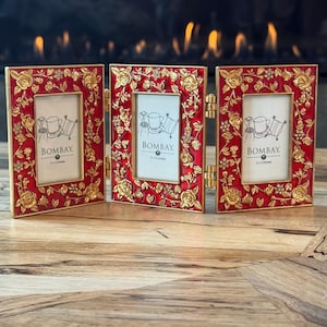 May include: A triptych photo frame featuring three rectangular panels connected by gold-tone metal hinges. Each frame has a vibrant red enamel border with raised gold-tone floral rose motifs and small clear crystals. Each insert reads BOMBAY 2x3 inches.