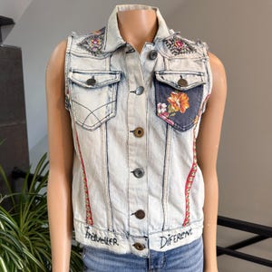 May include: A light-wash denim vest with a floral pocket design and embroidered details. The vest features decorative stitching, embellished shoulders, and the words "Traveller" and "Different" at the hem.