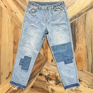 May include: Light blue denim jeans with distressed details and patches. The jeans feature a studded waistband and a relaxed fit. The hem is frayed, adding a casual touch. The patches are a darker shade of blue.