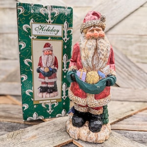 May include: A hand-painted Santa Claus figurine wearing a red coat with white trim, holding a green sack filled with gifts. Beside it stands a green cardboard box labeled Holiday Treasures with a picture of the same Santa Claus figurine.