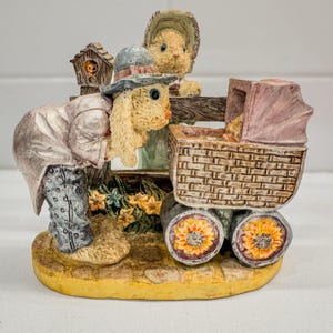 May include: A detailed figurine featuring two bunny figures, one leaning over a wooden fence and the other standing behind it. A baby carriage with sunflower wheels and a birdhouse are also present. The color palette includes beige, yellow, and pink.