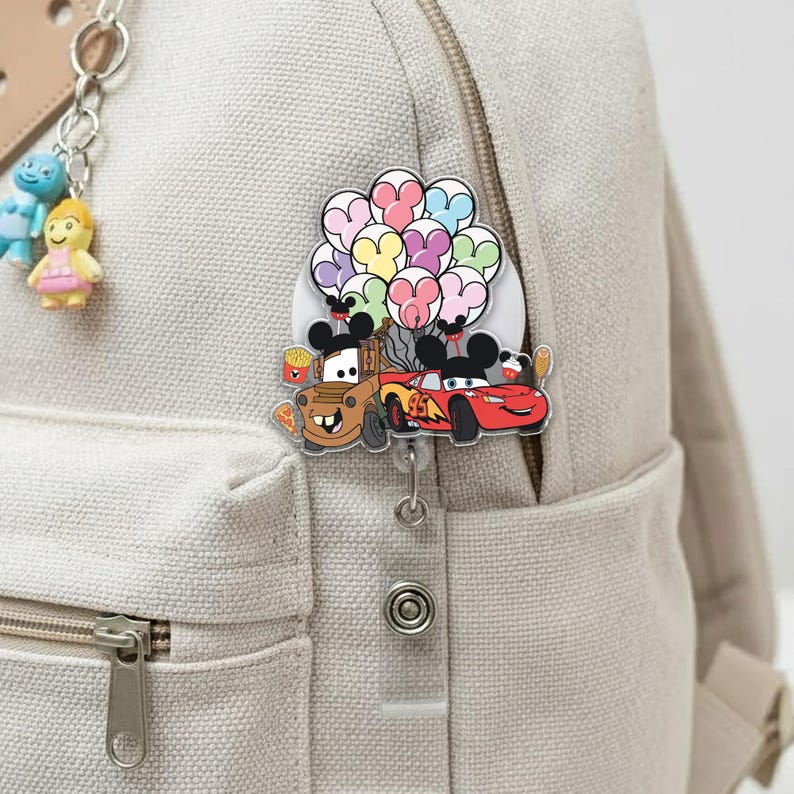 Op de afbeelding: Een intrekbare badge reel met cartoon auto's Mater en Lightning McQueen met Mickey Mouse oren, met kleurrijke ballonnen. De badge reel is bevestigd aan een beige rugzak, met een kleine sleutelhanger van cartoonfiguren.