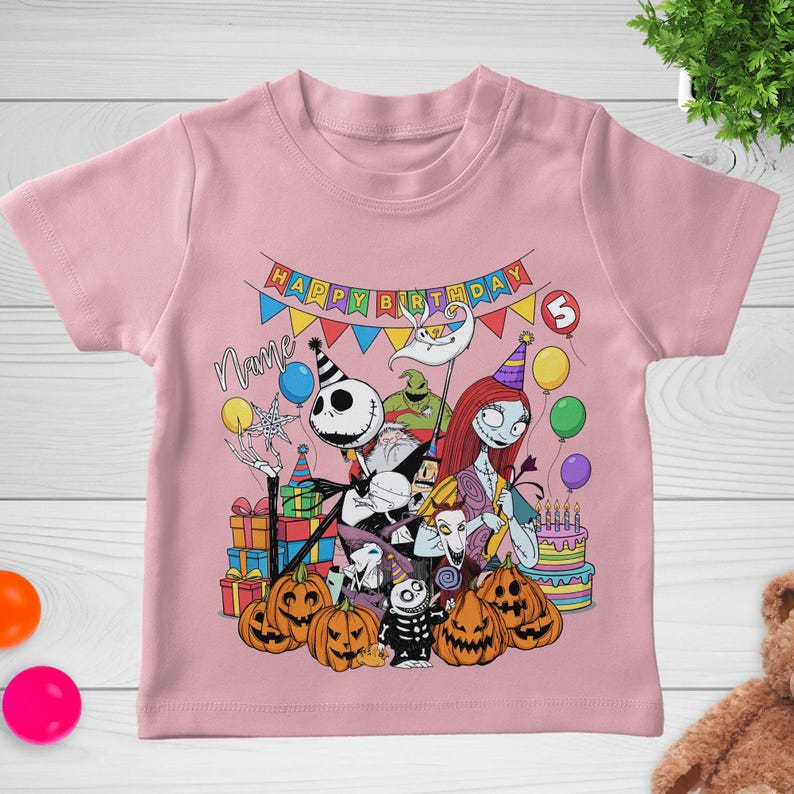 May include: Pink short-sleeved t-shirt featuring a colorful graphic of characters from The Nightmare Before Christmas. The design includes Jack Skellington, Sally, and pumpkins, with a "Happy Birthday" banner and a birthday cake.