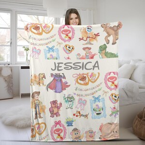 Puede incluir: Una manta color crema con varios personajes de Toy Story y elementos decorativos. La manta incluye el nombre "JESSICA" en letras grises grandes. El dise&ntilde;o incorpora marcos de dibujos animados, lazos y corazones, creando una est&eacute;tica l&uacute;dica y caprichosa.