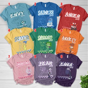 May include: A collection of nine colorful t-shirts, each featuring a different emotion from the movie "Inside Out." The shirts are in various colors, including blue, red, yellow, green, orange, pink, and purple, with the emotion's name and character illustration.