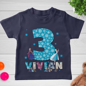 May include: Navy blue toddler t-shirt with a large number "3" filled with snowflakes. The name "VIVIAN" is printed below the number, with images of animated characters. The shirt is decorated with additional snowflake graphics.