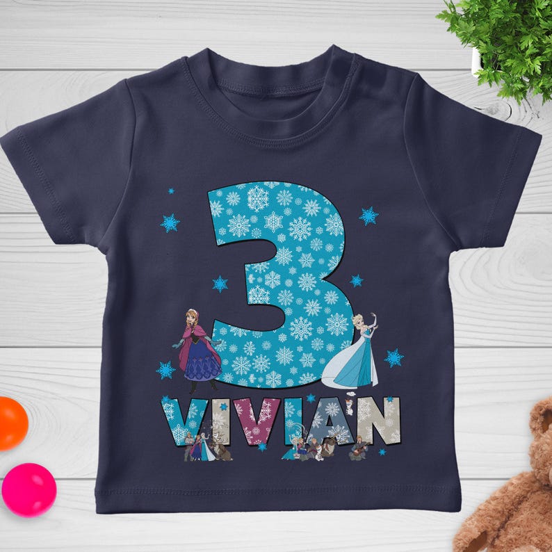 May include: Navy blue toddler t-shirt with a large number "3" filled with snowflakes. The name "VIVIAN" is printed below the number, with images of animated characters. The shirt is decorated with additional snowflake graphics.
