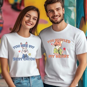 May include: Two white short-sleeved t-shirts featuring cartoon characters. The left shirt displays a rabbit with the text "YOU ARE MY BEST CATCH". The right shirt displays a fox with the text "YOU HUSTLED MY HEART" and a small red paw print.