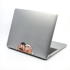 May include: A silver laptop with a sticker featuring cartoon Mr. and Mrs. Potato Head characters. The sticker shows the characters' faces with their distinctive features, including hats, mustaches, and smiles. The laptop is on a white surface.