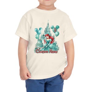 以下が含まれることがあります： プリンセス、お城、風船、ケーキのカラフルなグラフィックが特徴の、クリーム色の幼児用Tシャツ。風船には数字の3が表示されています。「Custom Name」の文字も表示されています。