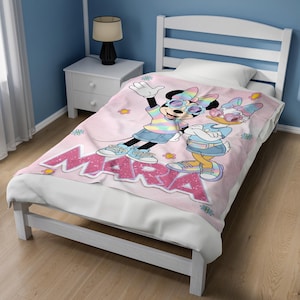 Op de afbeelding: Een roze deken met Minnie Mouse en Daisy Duck met een zonnebril en casual kleding. De deken heeft de naam "MARA" in grote, glinsterende roze letters. De personages staan tegen een achtergrond van kleine bloemen.