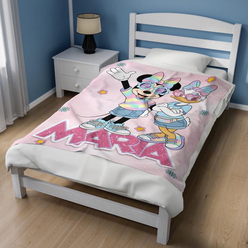 Op de afbeelding: Een roze deken met Minnie Mouse en Daisy Duck met een zonnebril en casual kleding. De deken heeft de naam "MARA" in grote, glinsterende roze letters. De personages staan tegen een achtergrond van kleine bloemen.