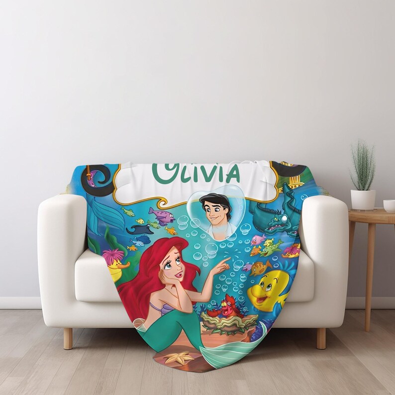 May include: A soft throw blanket featuring a colorful underwater scene with a red-haired mermaid, a yellow fish, a crab, and a man inside a heart-shaped bubble. The name OLIVIA is printed in large green letters at the top of the blue ocean background.