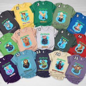 Op de afbeelding: Een collectie van vijftien t-shirts met korte mouwen in diverse kleuren, elk voorzien van een Zootopia-personage in een blauw schildembleem met de tekst Zootopia en de naam van het personage eronder gedrukt, genummerd van &eacute;&eacute;n tot vijftien.