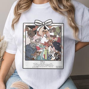 Puede incluir: Camiseta blanca con un gr&aacute;fico enmarcado de personajes de la pel&iacute;cula de animaci&oacute;n "Zootr&oacute;polis". El gr&aacute;fico incluye un lazo blanco en la parte superior y el t&iacute;tulo de la pel&iacute;cula y el a&ntilde;o 2026 en la parte inferior. El dise&ntilde;o es de estilo vintage.