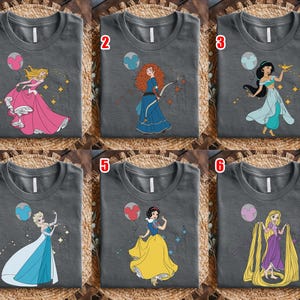 May include: Six charcoal gray t-shirts with different cartoon princess designs. Each shirt features a princess in a colorful dress with a matching balloon and sparkling accents. The princesses include Aurora, Merida, Jasmine, Elsa, Snow White, and Rapunzel.