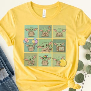 May include: Yellow t-shirt with a grid of nine illustrations of a green alien character in different poses. The character has large ears and a brown robe. The shirt has a crew neck and short sleeves.