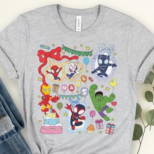 May include: A light gray t-shirt featuring a colorful graphic print of various superheroes including Iron Man, Hulk, and Spider-Man characters. The design includes party elements like balloons, a cake, gifts, and the text NAME 5 in balloons.