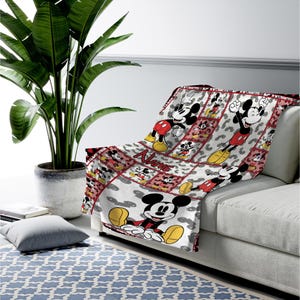 May include: A white and red Mickey Mouse throw blanket draped over a sofa. The blanket features a repeating pattern of Mickey Mouse in various poses, with a red border and the name "Name" printed on it. The blanket is made of a soft material.