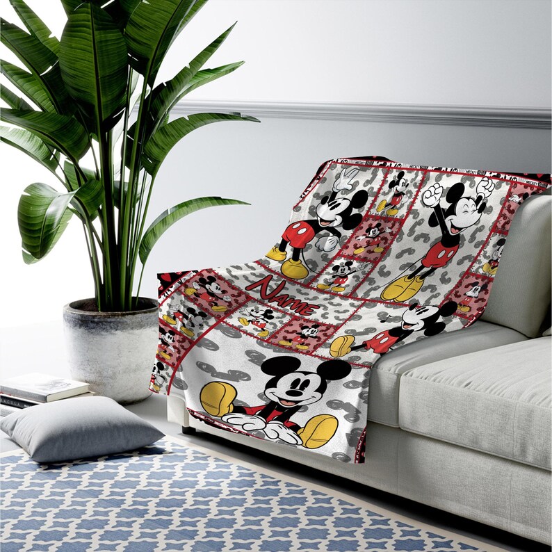 May include: A white and red Mickey Mouse throw blanket draped over a sofa. The blanket features a repeating pattern of Mickey Mouse in various poses, with a red border and the name "Name" printed on it. The blanket is made of a soft material.