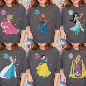 May include: Six dark gray t-shirts with different princess designs. Each shirt features a colorful illustration of a Disney princess, including Aurora, Merida, Jasmine, Elsa, Snow White, and Rapunzel, with a matching balloon.