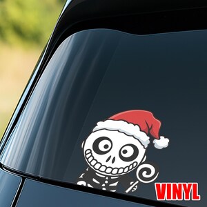 Op de afbeelding: Een wit en zwart vinyl sticker van een skelet karakter met een kerstmuts en een lolly. De sticker zit op een autoraam, met het woord "VINYL" in het rood.