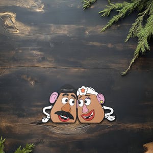 May include: Two cartoon potato head cutouts, one with a black hat and mustache, the other with a white headband and flower. Both have large eyes, pink ears, and red smiling mouths. They are set against a dark wood background with green sprigs.
