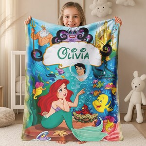 May include: A rectangular fleece blanket featuring a vibrant underwater scene with the name Olivia printed in the center. The design includes characters like Ariel, King Triton, Ursula, Flounder, and Prince Eric, set against a blue ocean background.