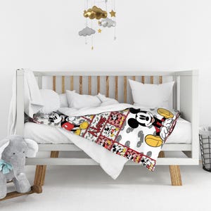 May include: A white baby crib with a colorful blanket featuring Mickey Mouse. The blanket has a patchwork design with Mickey Mouse illustrations in red, black, yellow, and white. A gray elephant rocking toy is in the foreground.