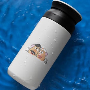 May include: A white insulated tumbler with a black lid, featuring a sticker of two cartoon potato characters. The tumbler is cylindrical and appears to be made of stainless steel. The background is a blue surface with water droplets.