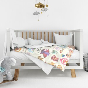 Puede incluir: Una manta de beb&eacute; color crema con ilustraciones de personajes de dibujos animados coloridos, incluyendo una figura con t&uacute;nica p&uacute;rpura, un oso de peluche rosa y un extraterrestre verde. La manta est&aacute; sobre una cuna blanca.