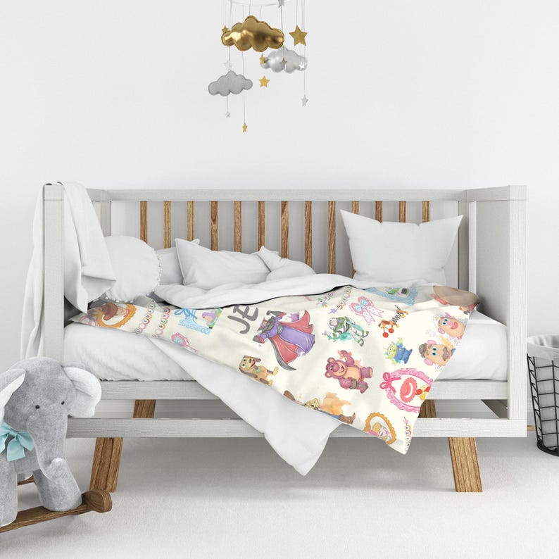 Puede incluir: Una manta de beb&eacute; color crema con ilustraciones de personajes de dibujos animados coloridos, incluyendo una figura con t&uacute;nica p&uacute;rpura, un oso de peluche rosa y un extraterrestre verde. La manta est&aacute; sobre una cuna blanca.