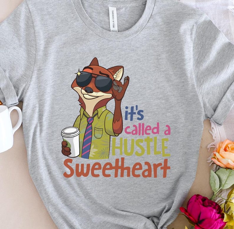 Op de afbeelding: Een lichtgrijs t-shirt met een cartoonvos met een zonnebril en een koffiekopje. De vos draagt een groen overhemd en een stropdas. De tekst op het shirt luidt "it's called a HUSTLE Sweetheart."